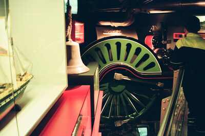 A green locomotive driving wheel beside a ship model — London, England (August 2000)